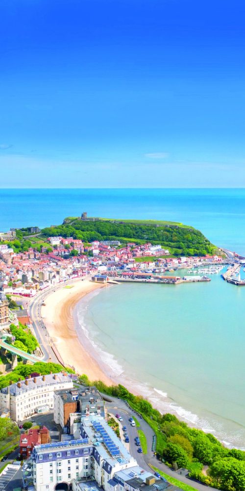 Aerial photo of the town centre of Scarborough in East Yorkshire in the UK showing the coastal beach and harbour with boats and the Scarborough Castle on a bright sunny summers day